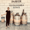 Smiling woman in black stands with arms wide beside three wooden wine barrels and bottles at Parker Coonawarra Estate sign.
