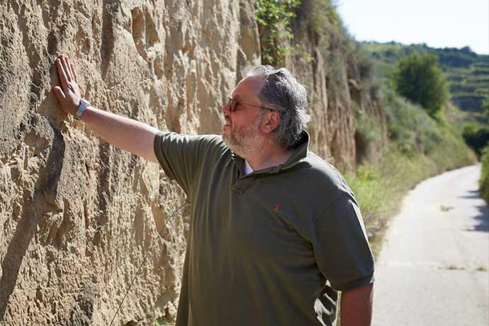 Older man in a green polo shirt rests his hand on a rough stone wall along a rural road, enjoying a sunny day.