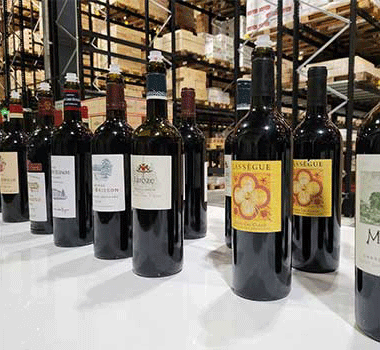row of red wine bottles lined up on a white surface with warehouse-style shelves in the background (wine shop).